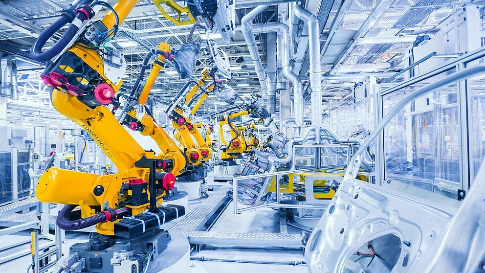 Blick in eine Produktionshalle der Automobilindustrie. Eine Reihe Roboterarme schraubt an Blechteilen.